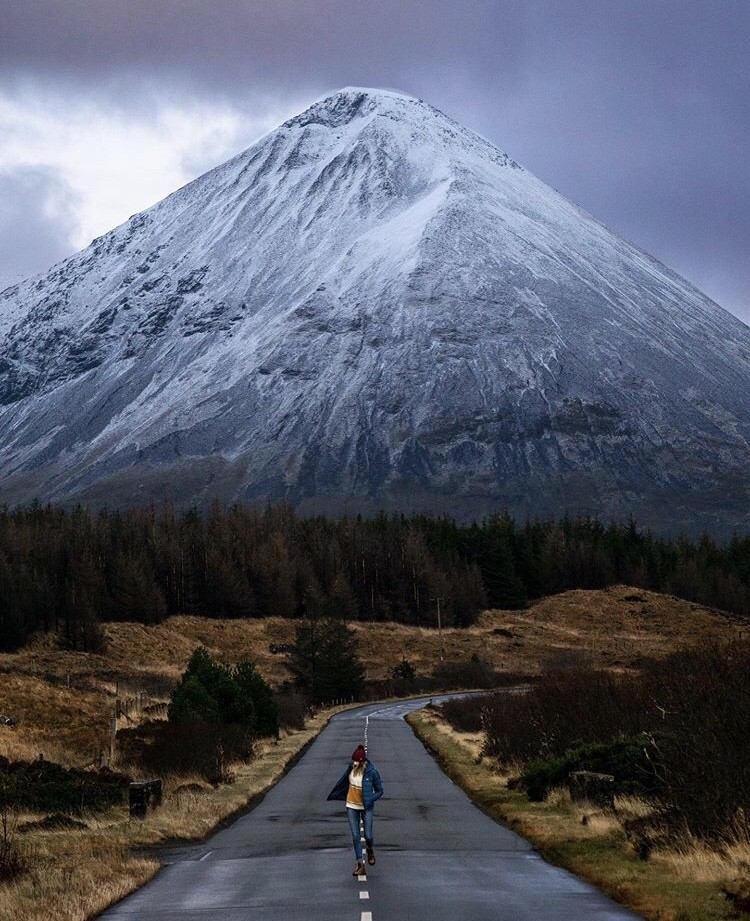 A lady walking in the middle of the road in front of a snow capped mountain on a trip to Scotland